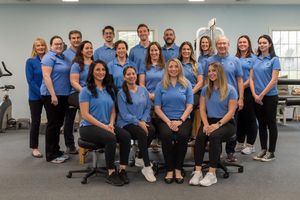 A group of healthcare professionals in matching blue polo shirts posing together in a modern physical therapy clinic.