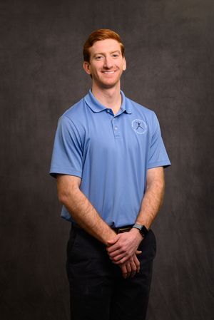 Young man with red hair smiling in a light blue polo shirt with a logo, posing against a gray backdrop.