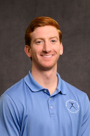 Smiling man with red hair wearing a light blue physical therapy sports polo shirt against a gray background.