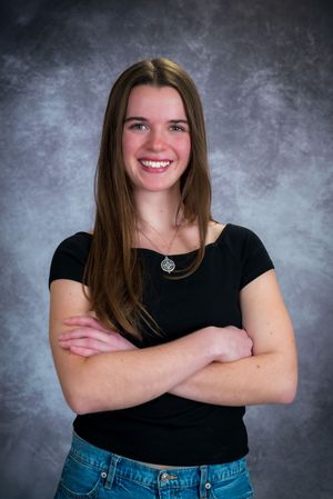Smiling young woman with long brown hair, arms crossed, wearing black top and jeans, posing for portrait photo.
