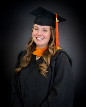 Smiling graduate in black cap and gown with orange tassel and stole poses for professional portrait photo.