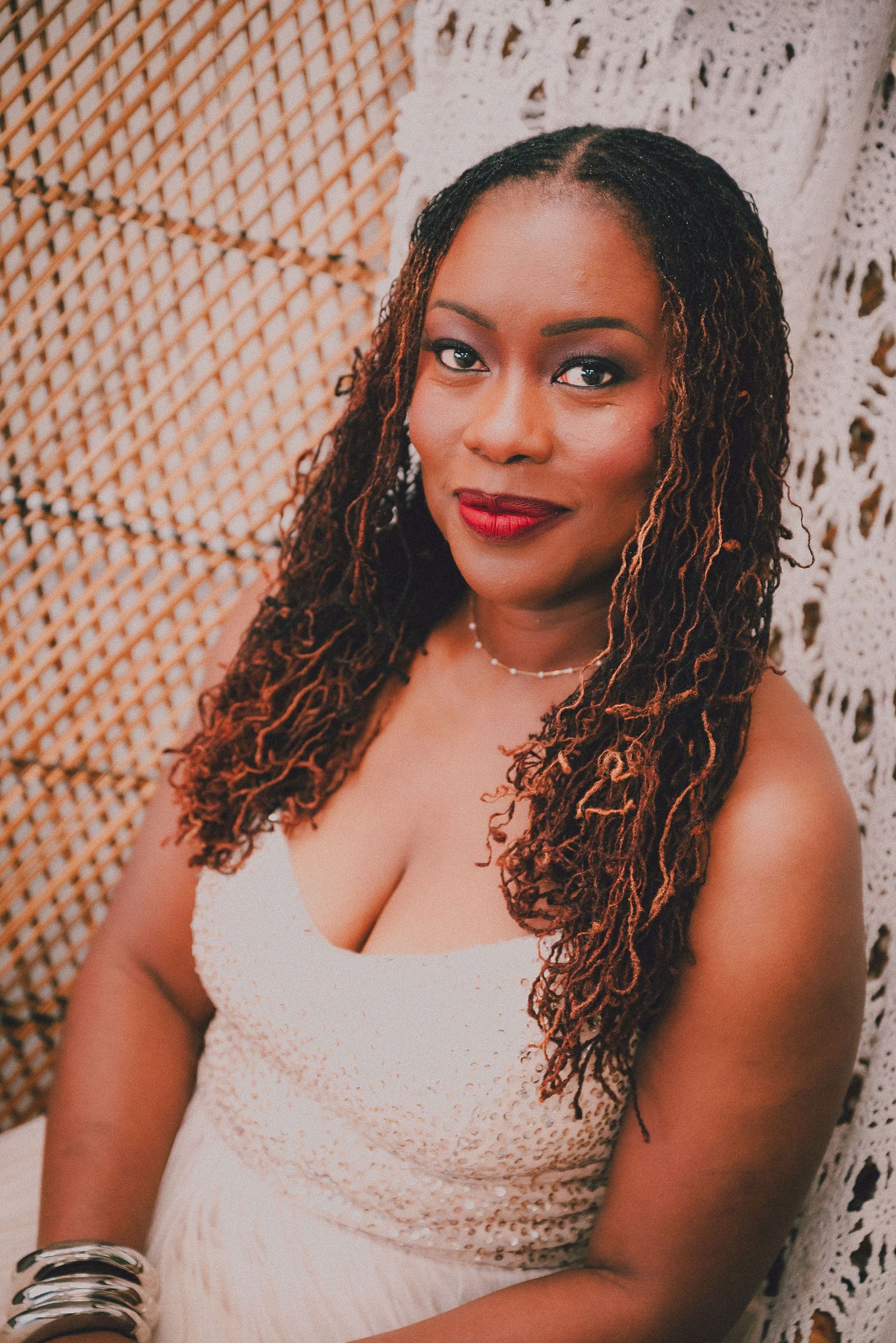 Woman with curly locs wearing white dress and red lipstick posing against woven backdrop.