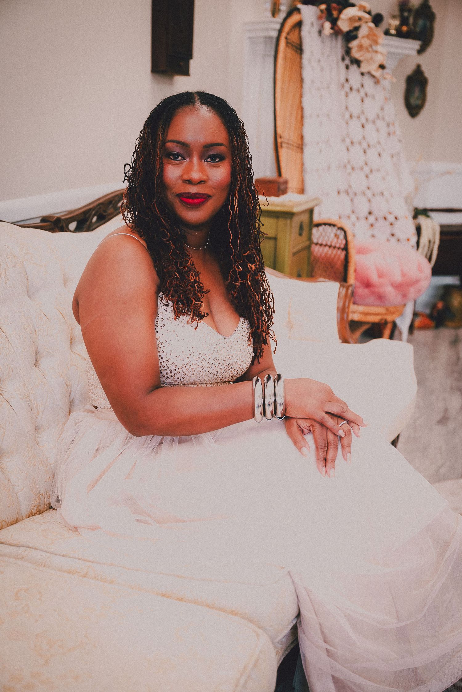 Woman in white beaded gown sitting elegantly on ivory sofa with red lipstick and silver bracelet.