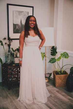 Woman in elegant white lace wedding gown smiling in a decorated room with plants and artwork.