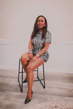 Smiling woman in silver sequin romper and black heels sitting on a stool in a bright studio setting.