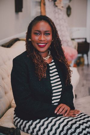 Professional woman in black blazer and striped outfit smiling confidently while seated on a white sofa indoors.