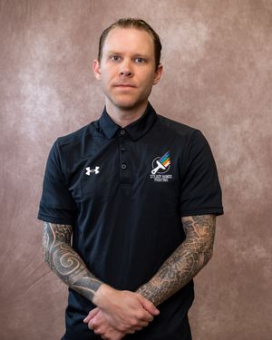 Man in black Under Armour polo with colorful logo and sleeve tattoos poses against brown studio backdrop.