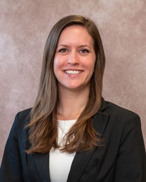 Professional headshot of a smiling woman with long brown hair wearing a black blazer and white blouse.