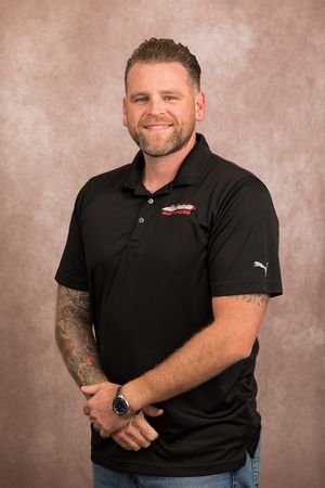 Smiling man with tattoos wearing a black Puma polo shirt with a logo, posing for a professional headshot.
