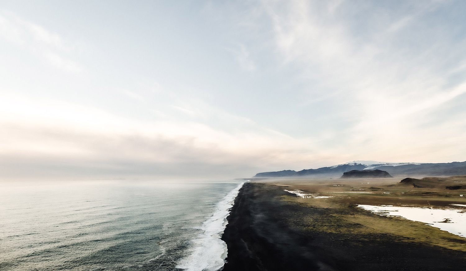 Aerial view of Iceland's black sand beach coastline with dramatic ocean waves, snow-capped mountains, and misty sky.