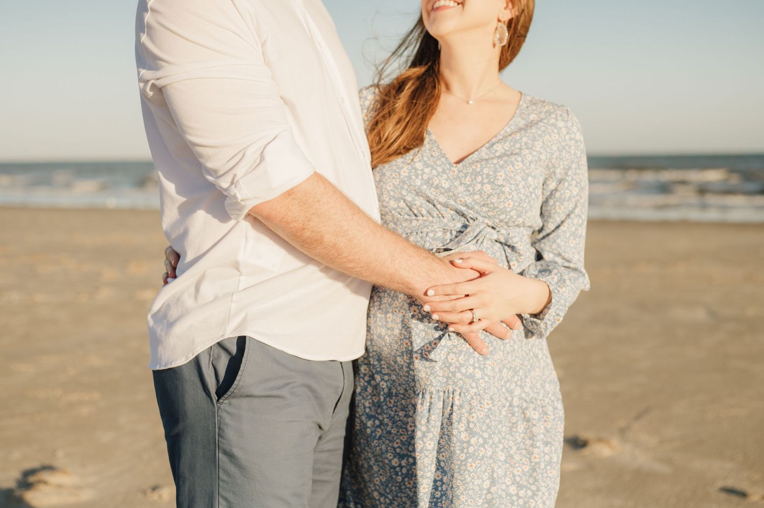 Couple in casual attire embracing on a sunny beach with ocean waves in the background.