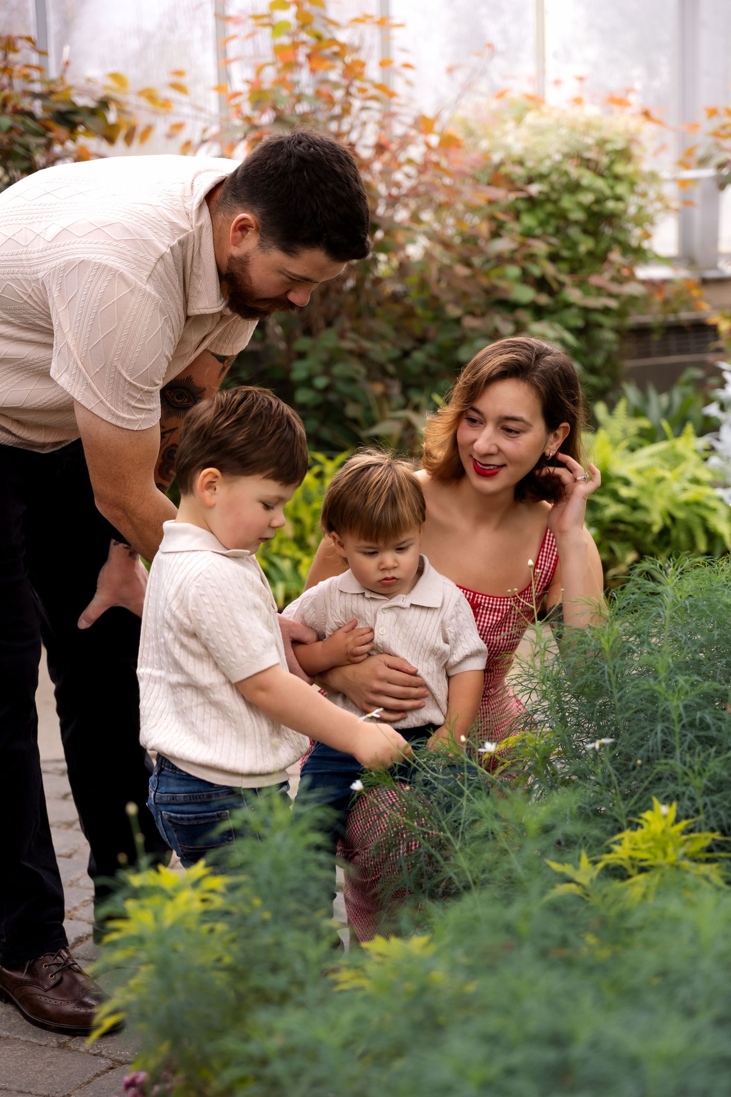 Young family with two toddler boys exploring green plants together in a lush greenhouse.