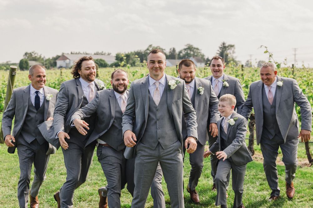 Groomsmen in grey suits celebrate with enthusiasm in outdoor vineyard setting.