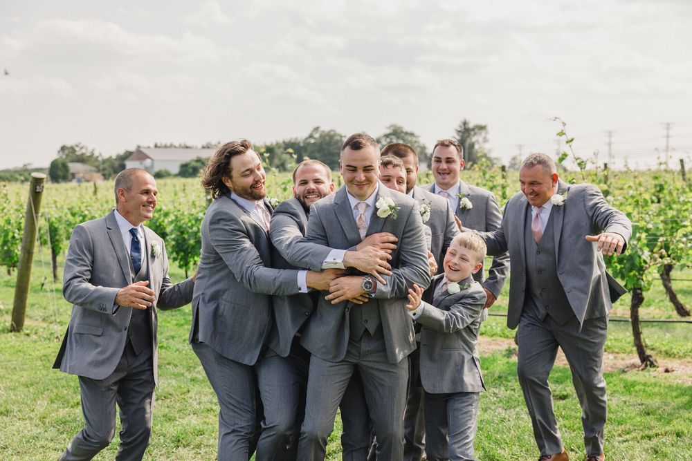 A group in gray suits laughing and celebrating together in a vineyard setting on a cloudy day.