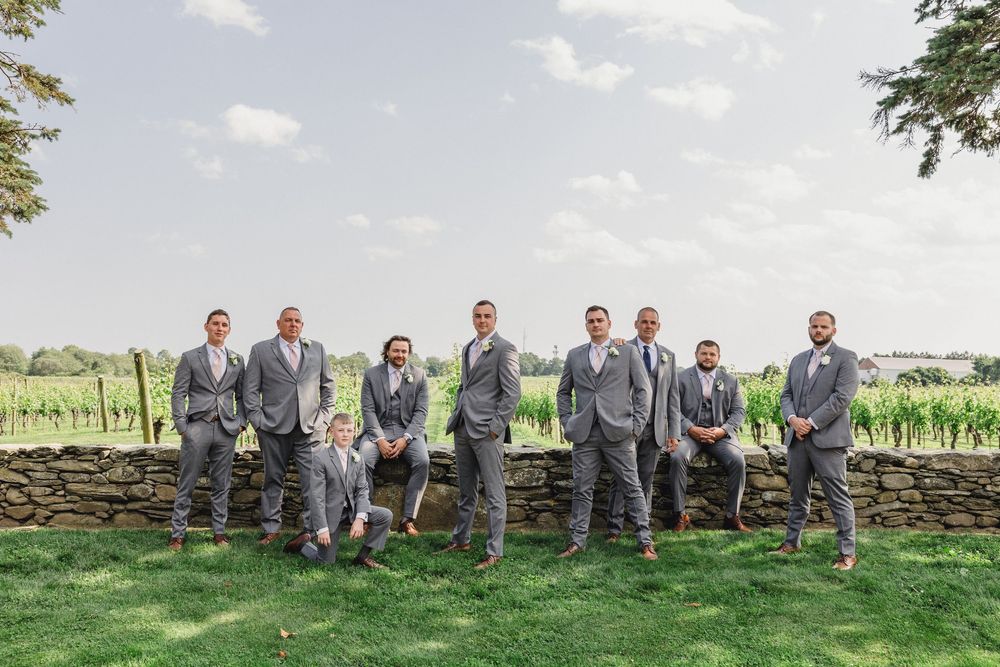 Wedding party in grey suits poses casually on a stone wall with vineyard backdrop.