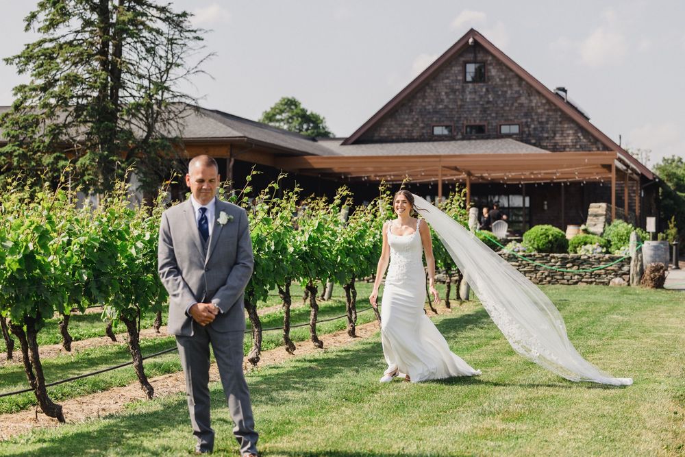 A wedding couple poses in front of a rustic barn surrounded by vineyard rows.