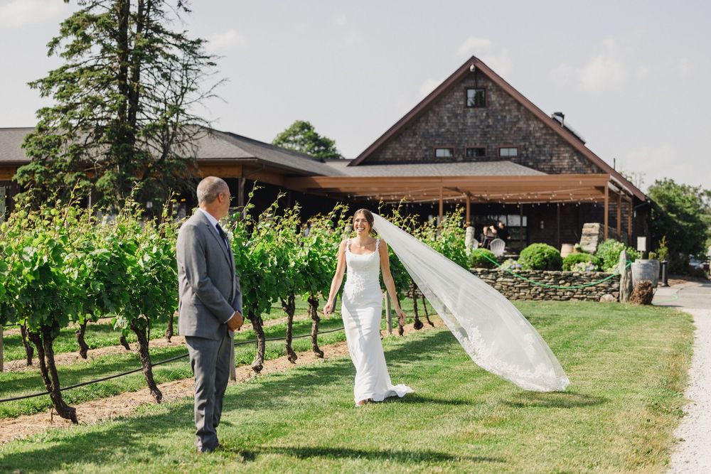 A couple in wedding attire stands in front of a rustic barn with a flowing veil caught in the breeze.