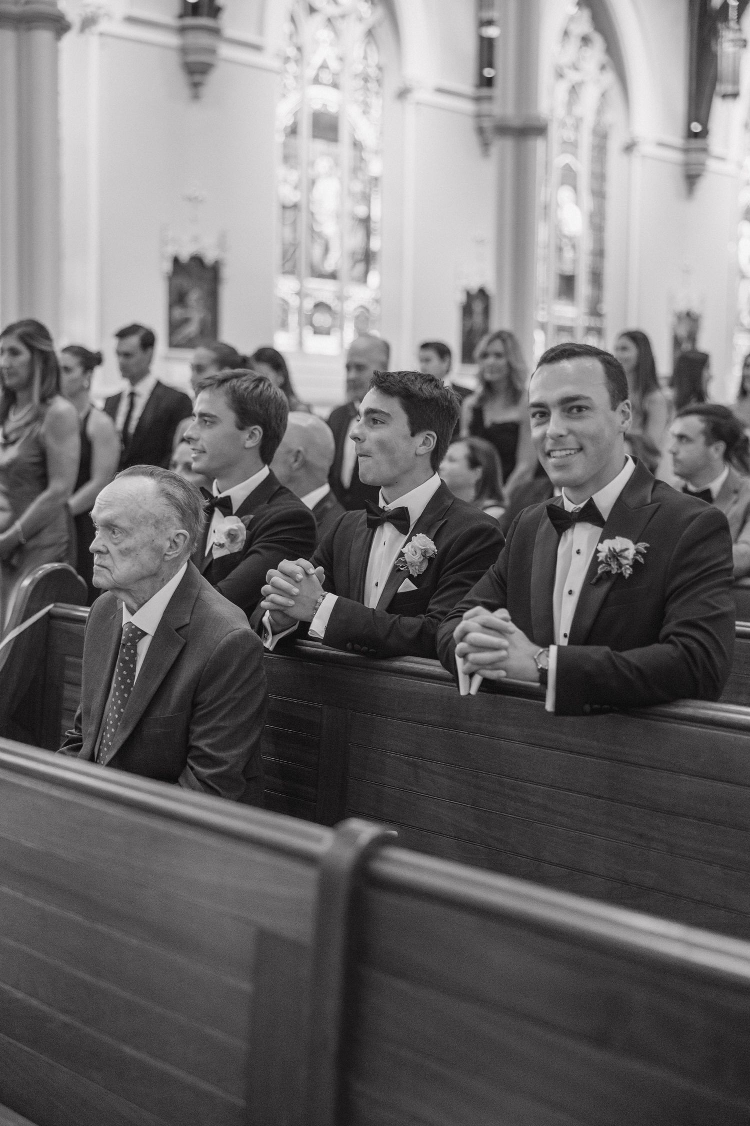 Black and white photo of wedding guests sitting in church pews smiling and laughing during ceremony.