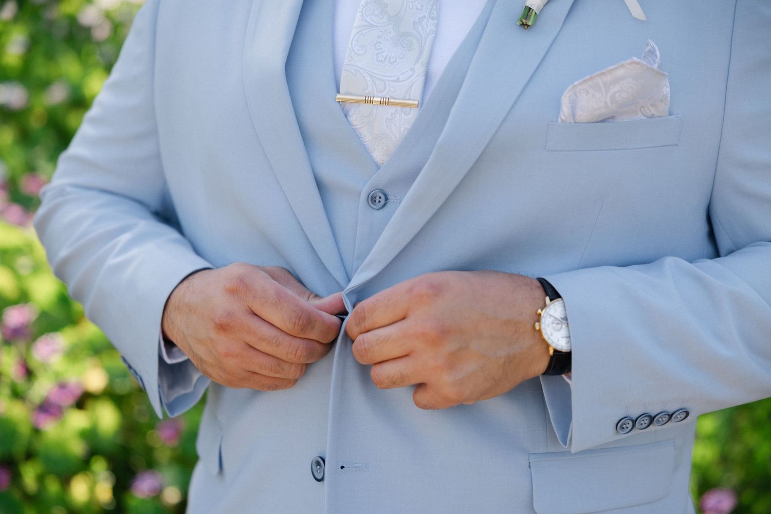 A person in a light gray suit poses outdoors on a path surrounded by trees and flowering bushes in a garden setting.