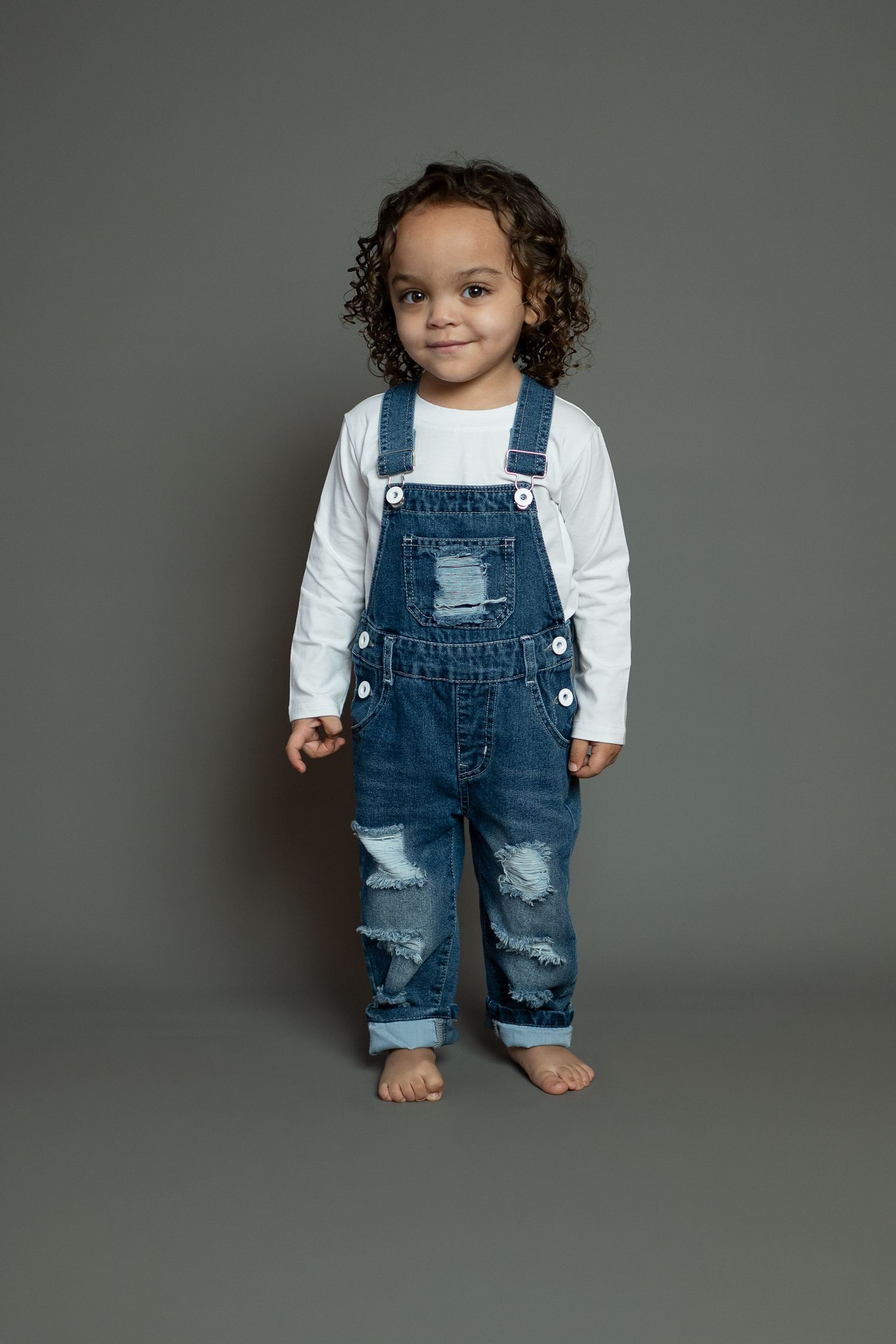 Small child wearing distressed denim overalls and white long sleeve shirt poses barefoot against gray backdrop.