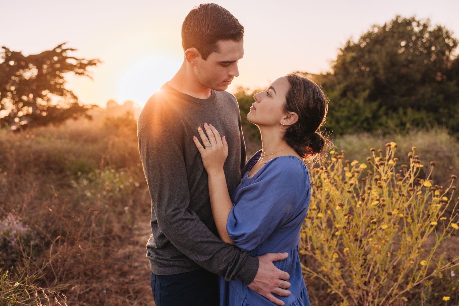 A romantic couple embraces in a golden sunset with flowering yellow bushes in the background.