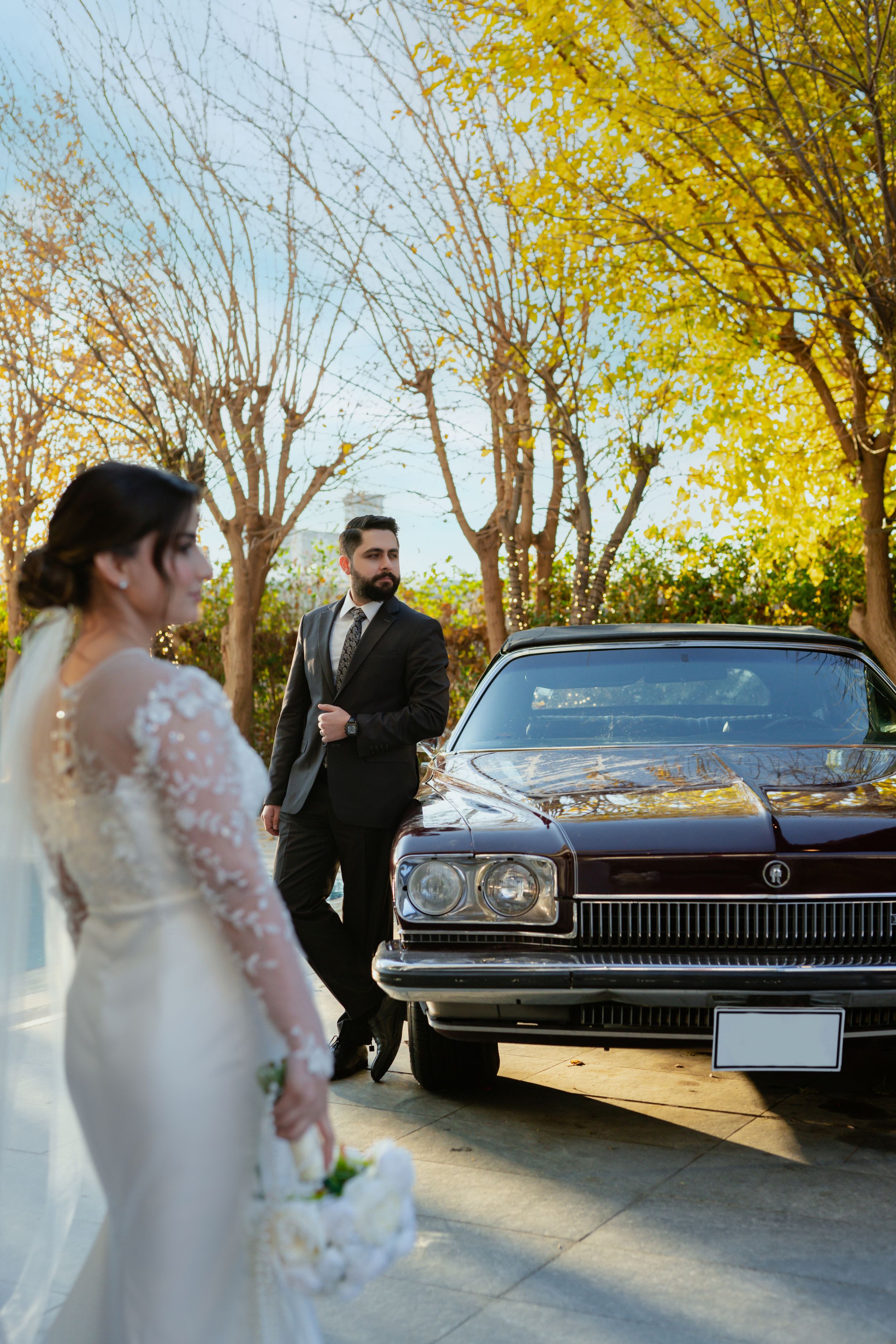 A classic maroon car parked under autumn trees at an outdoor wedding.