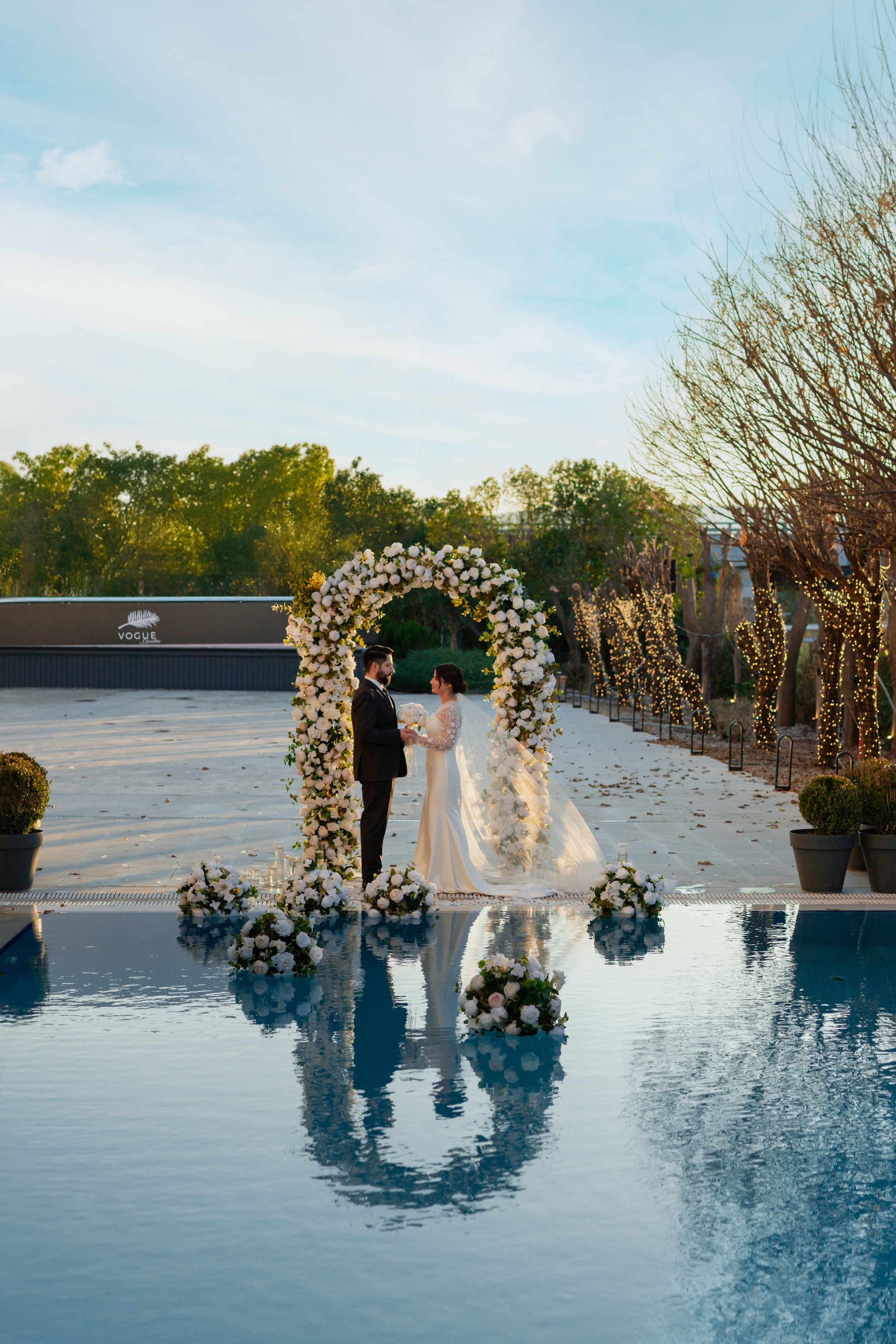 A romantic wedding ceremony setup with a floral arch and its reflection in a serene pool under a blue sky.