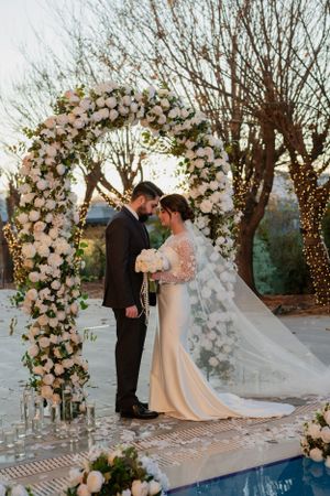Romantic wedding ceremony with couple standing beneath a grand floral arch with white roses and twinkle lights at dusk by a pool.
