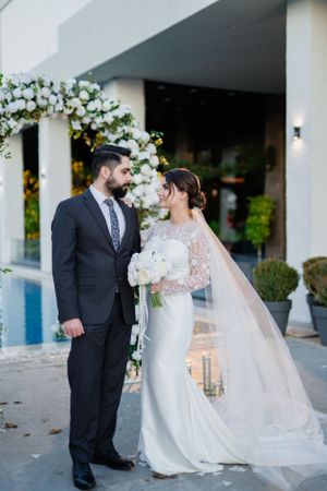 A romantic wedding couple stands by a pool under a white floral arch with flowing veil and elegant formal attire.
