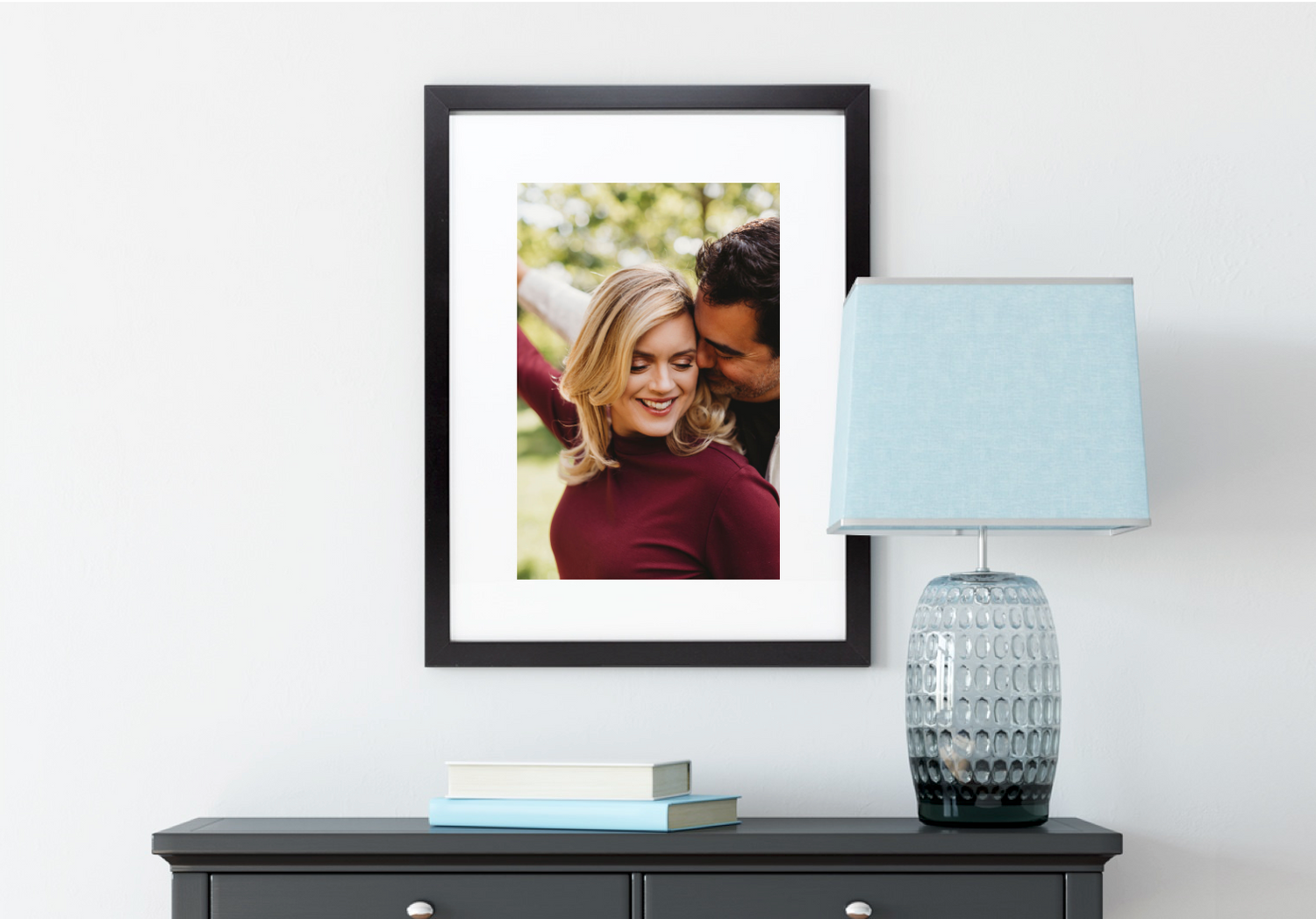 Framed photograph displayed above dark dresser with modern blue table lamp and decorative glass vase.