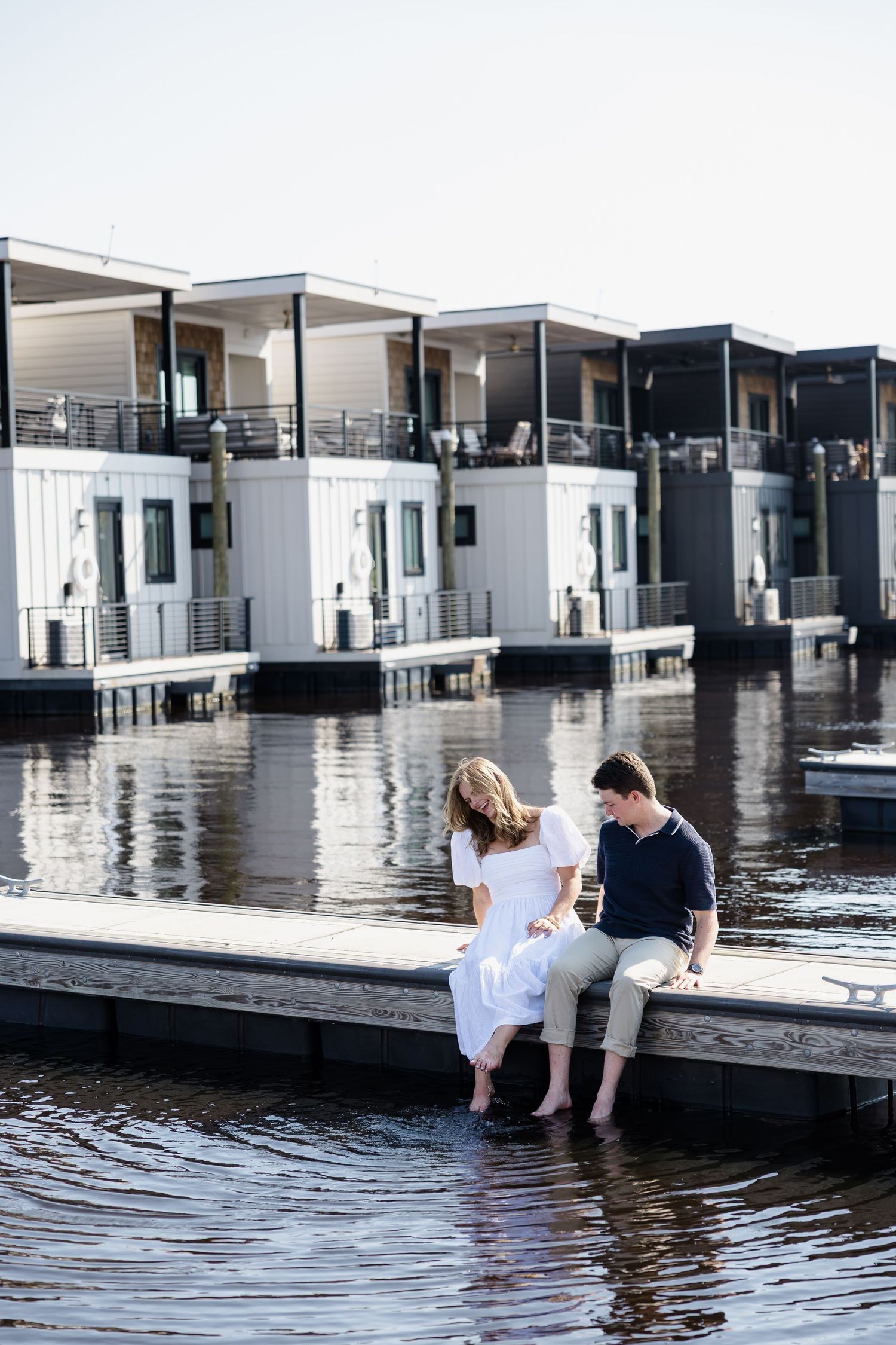 Two people dangle their feet in reflective water by modern white buildings on a sunny day.