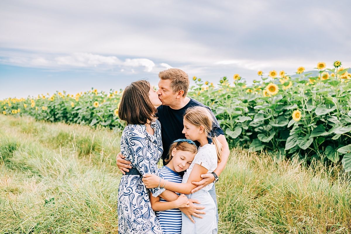 Family of 4 mother and father kissing and two younger daughters are hugging parents Canton Vaud Switzerland in front of a sunflower field
