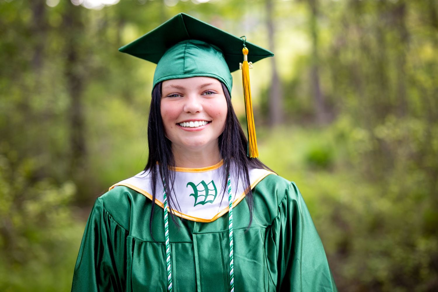 Graduate in emerald green cap and gown smiles for a portrait in an outdoor wooded setting.