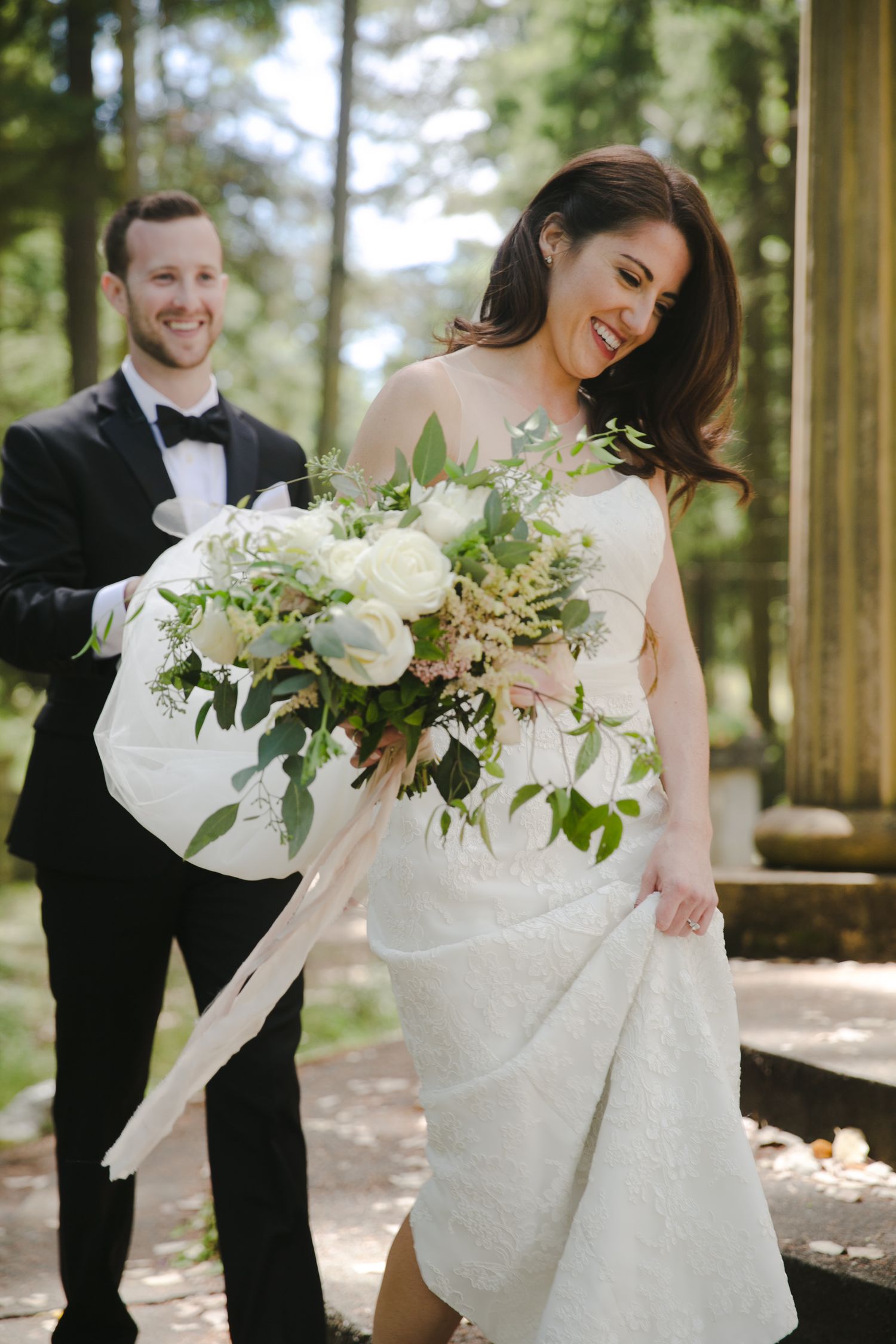 A joyful wedding moment outdoors showing a couple laughing together with a stunning white floral bouquet.