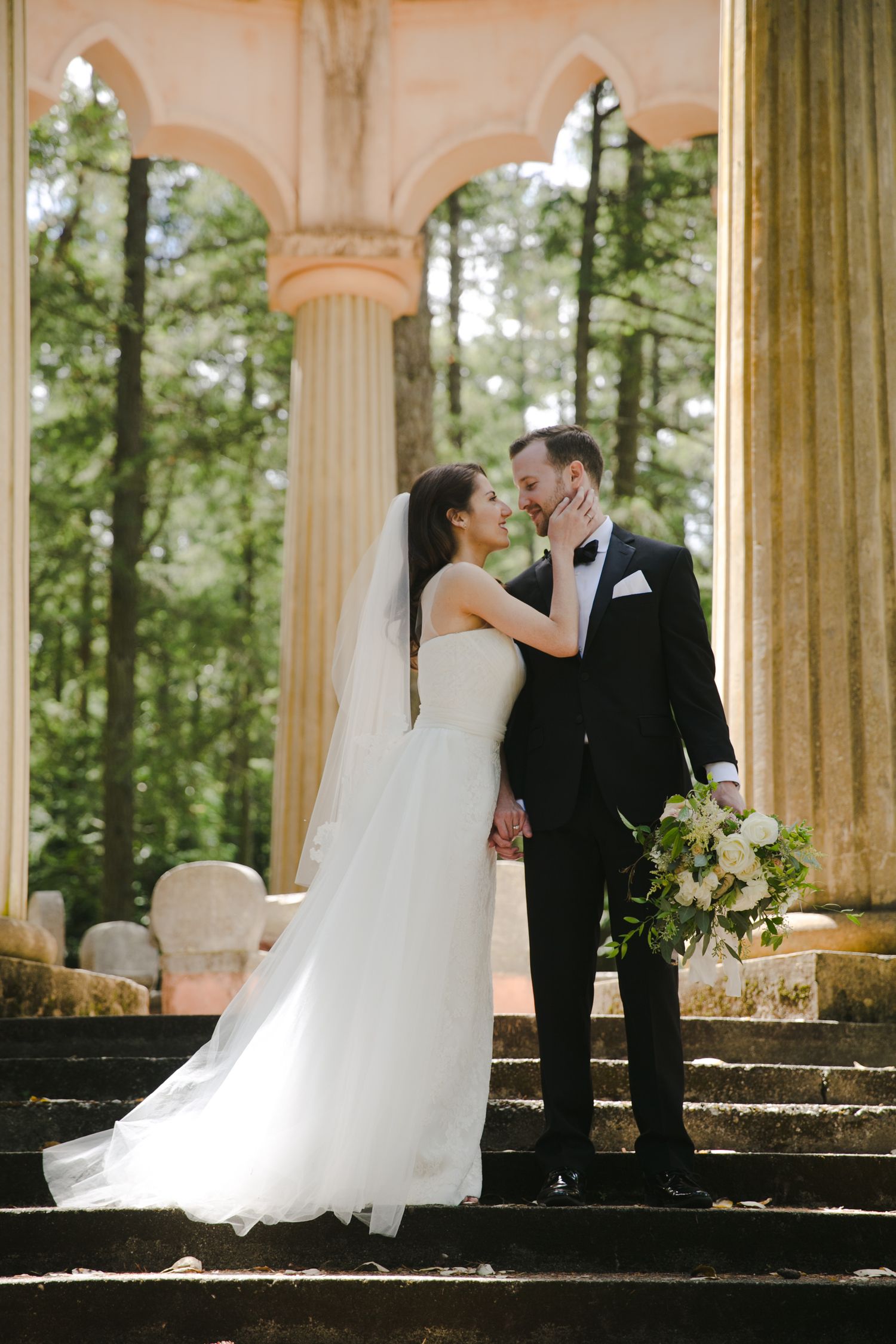 A bride and groom share an intimate moment on stone steps framed by elegant columns and greenery in the background.