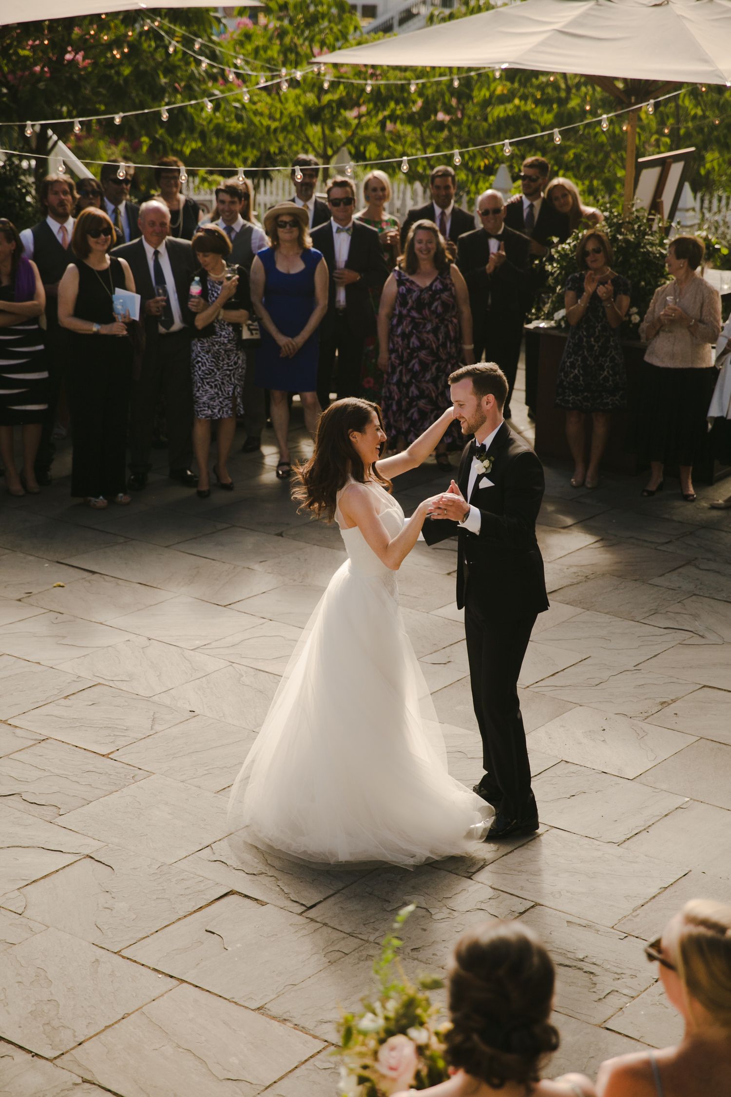 A couple shares their first dance at an outdoor wedding reception while guests watch from the stone patio under string lights.
