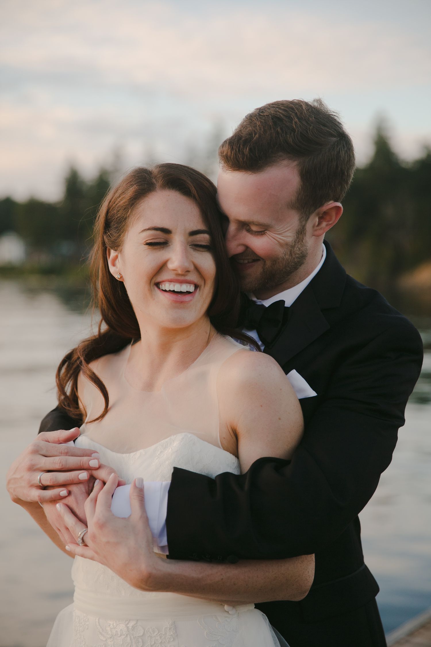 Joyful couple embraces in formal wedding attire by the water during sunset with evergreen trees in background.