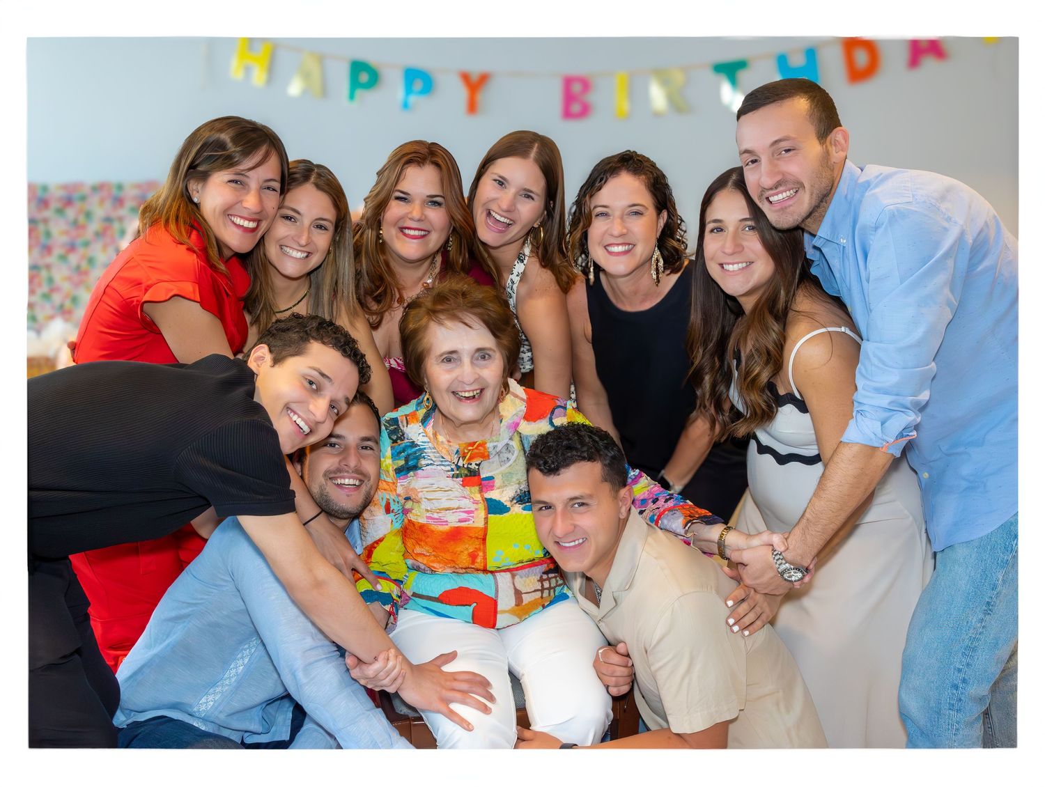 Birthday celebration group photo with colorful banner and cake surrounded by friends.
