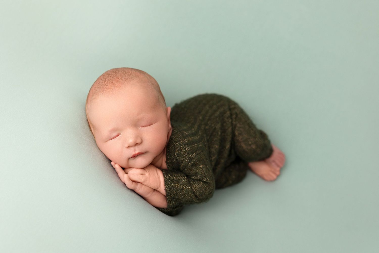Sleeping newborn wrapped in brown knit blanket on mint green background.