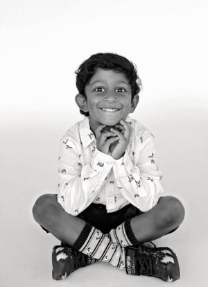 Black and white photo of child sitting cross-legged with hands under chin and cheerful expression.