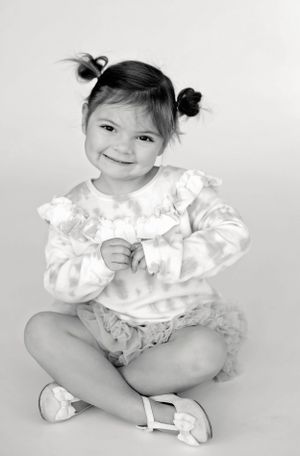 A toddler in ruffled white clothing sits cross-legged with pigtails against a white background.