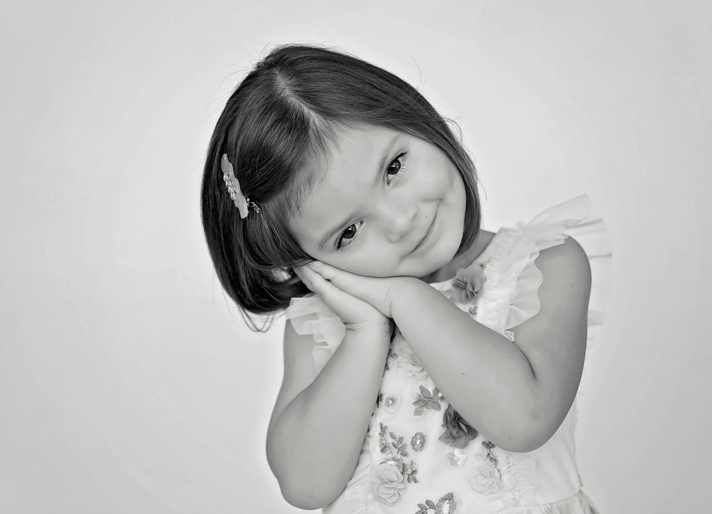 Black and white headshot of child with hands clasped near cheeks making sweet expression.
