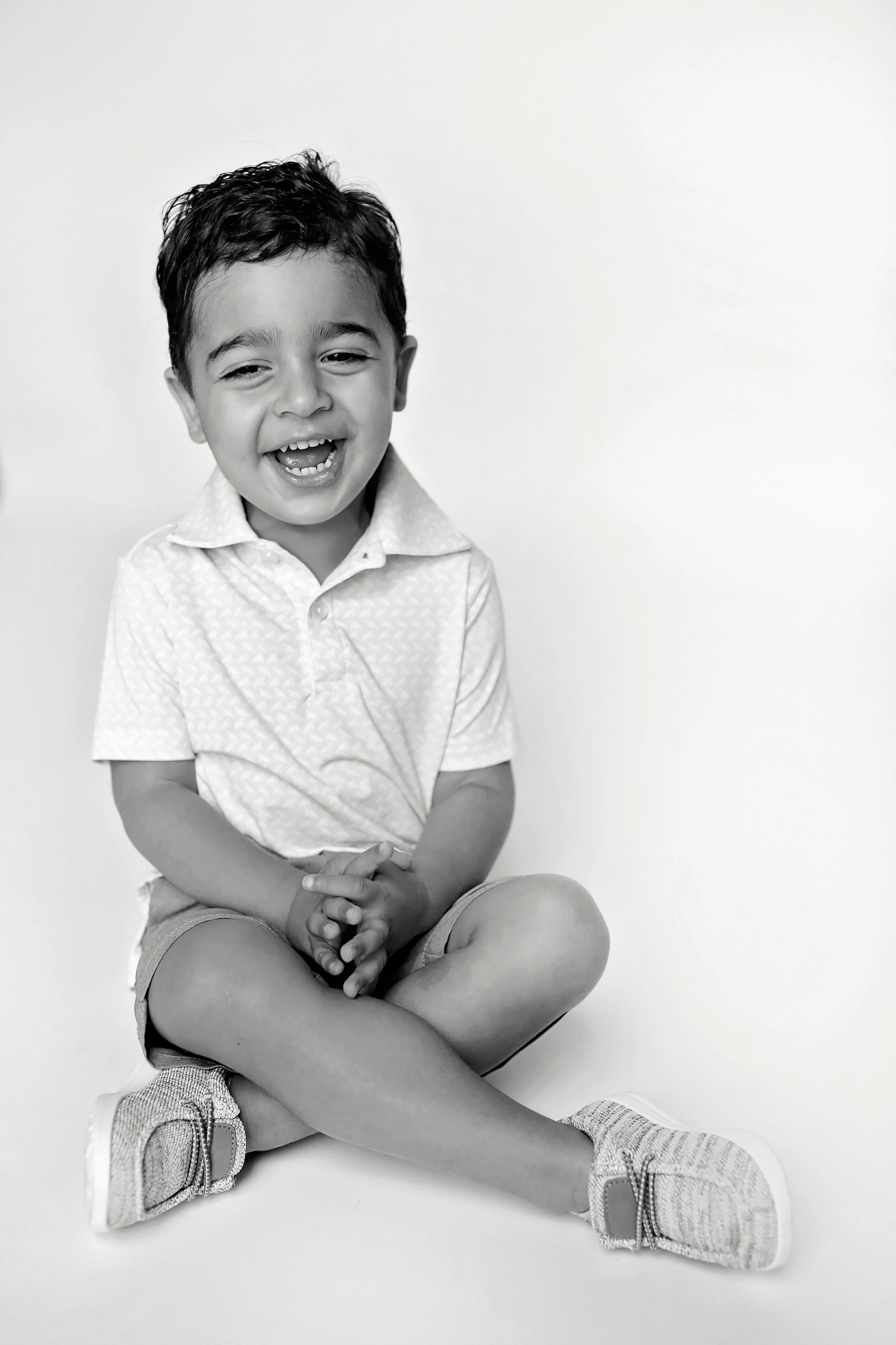 Black and white portrait of child sitting cross-legged in white polo shirt with happy expression.