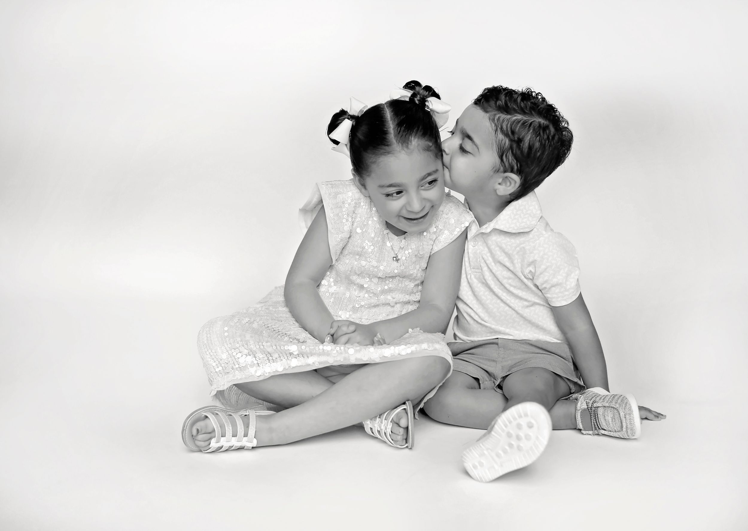 Black and white portrait of two children sitting together with one giving the other a kiss on cheek.