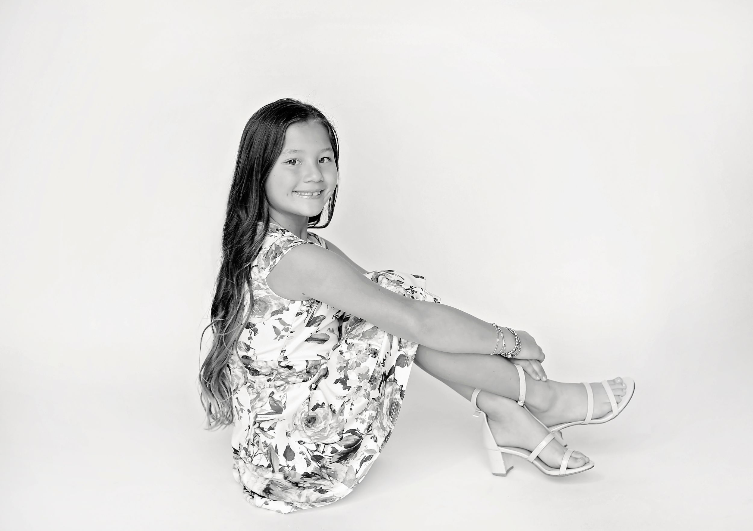 A black and white portrait of a young person in a floral dress sitting against a white background.