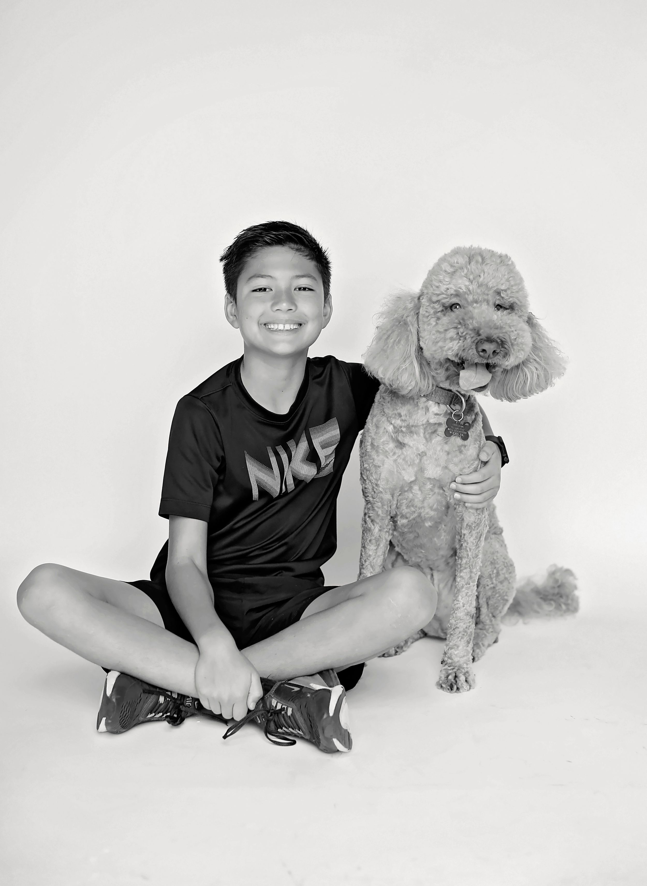 A black and white photo of a child and a poodle dog sitting together against a plain backdrop.