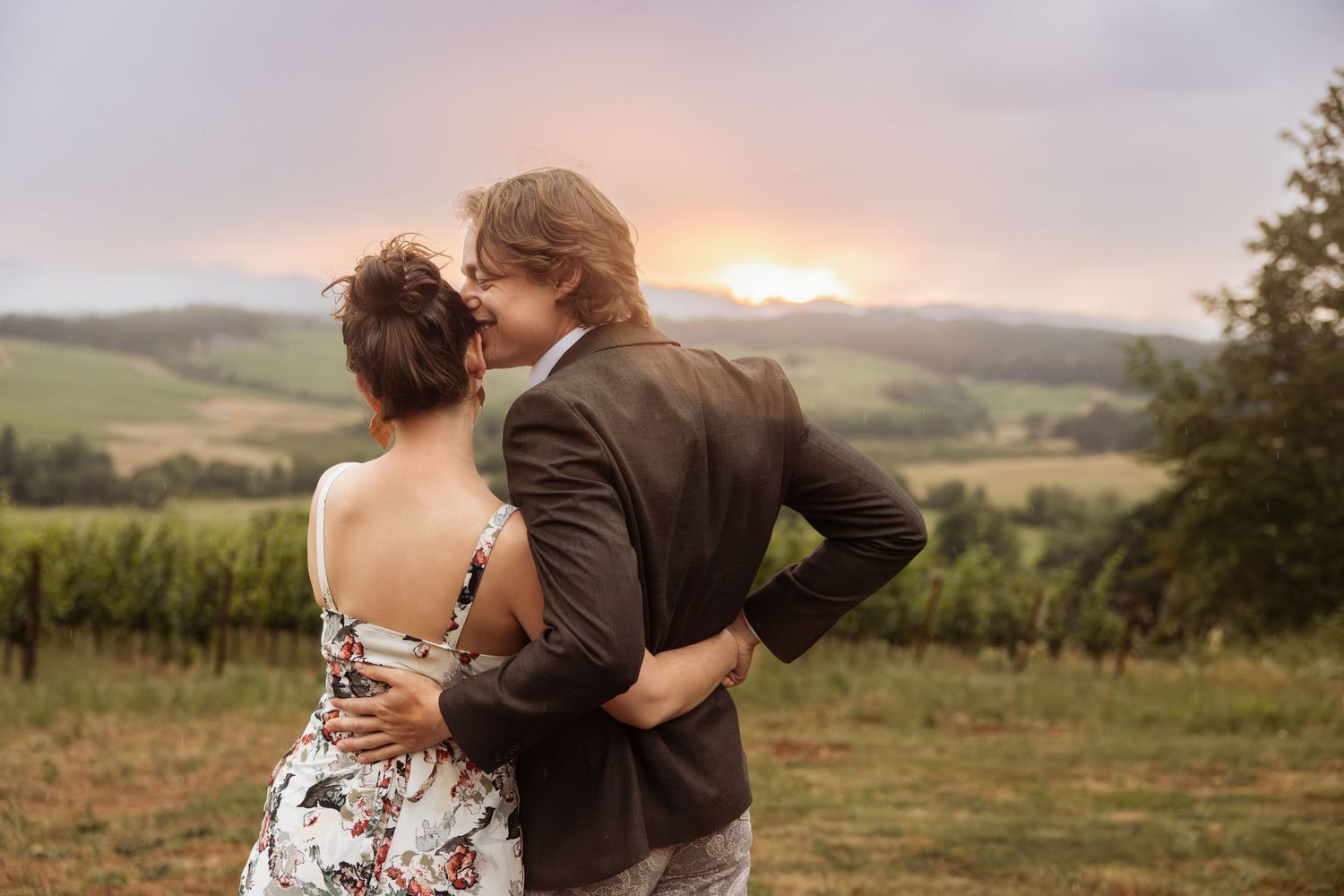 A couple embraces while watching sunset over rolling vineyard hills in an idyllic countryside setting.