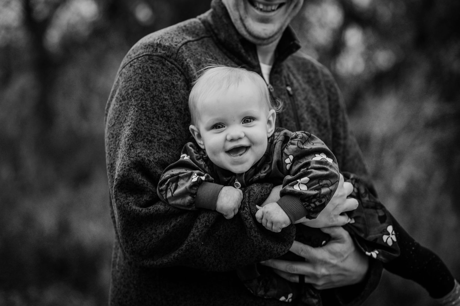 A black and white portrait of a baby wearing camouflage print being held and smiling outdoors.