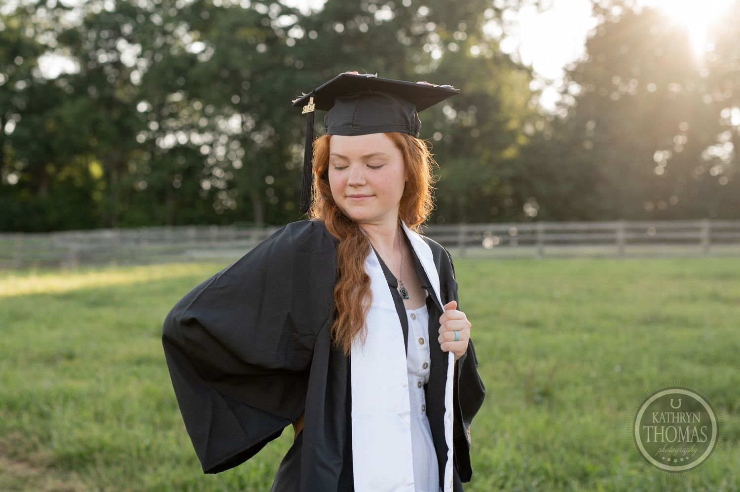 Graduate poses with a bright smile in graduation cap and gown at sunset in a grassy field.