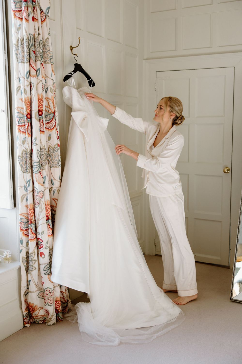 Wedding dress being prepared on a hanger against floral curtains in bright room.